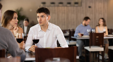 Three friends meeting in modern restaurant - men and a woman drinking wine, talking, discussing problems