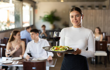 Young female waitress posing with tray of salad and wine in restaurant..
