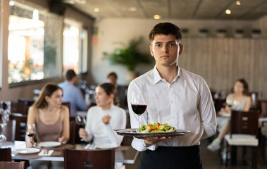 Young male waiter in uniform posing with tray and glass of red wine in his hands in restaurant