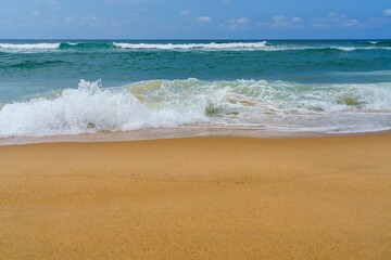 The image features a sandy beach with gentle waves under a clear blue sky. The soft sand and clear water create a perfect setting for swimming or relaxing while enjoying the view.