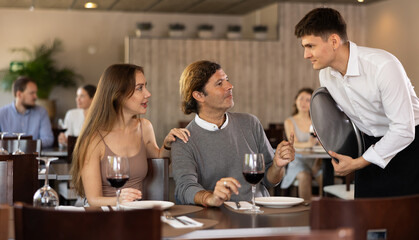 Young guy waiter taking order from couple adult man and young woman in restaurant