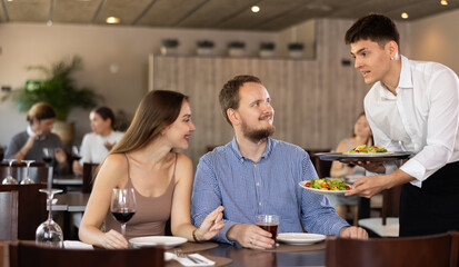 Young guy waiter brings order from couple young man and young woman in restaurant