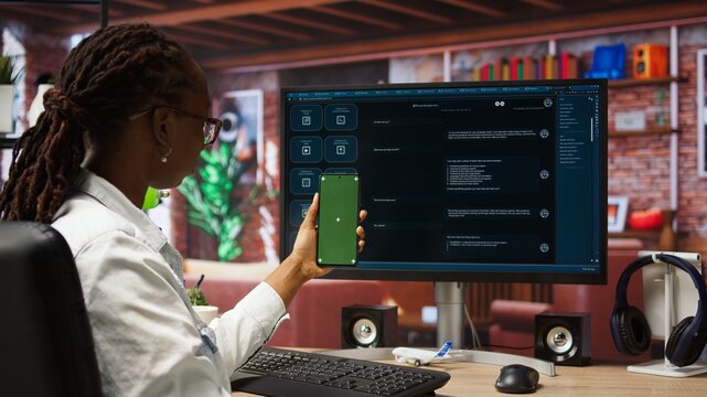 Woman seated at desk using AI LLM chatbot software and green screen phone in modern urban home office. Person uses large language model program on computer and mockup smartphone in apartment, camera A