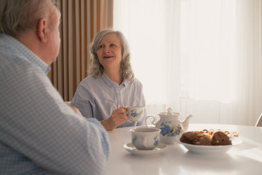 An elderly couple sits at a bright table, enjoying tea and conversation, showcasing warmth and companionship in a peaceful home environment, surrounded by beautifully crafted tableware and sunlight.