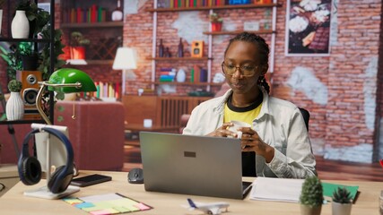 Remote worker at home office desk drinking coffee and browsing internet on notebook, researching information. Woman enjoying hot beverage while using search engines to find data for work, camera B