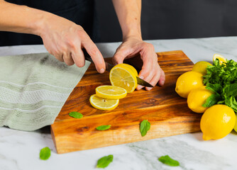 Close-up of hands slicing fresh lemons on a wooden cutting board with a bundle of whole lemons, mint leaves, and a green towel on a marble surface.