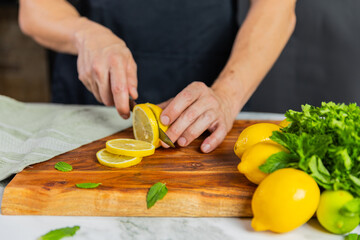 Close-up of hands slicing fresh lemons on a wooden cutting board with a bundle of whole lemons, mint leaves, and a green towel on a marble surface.