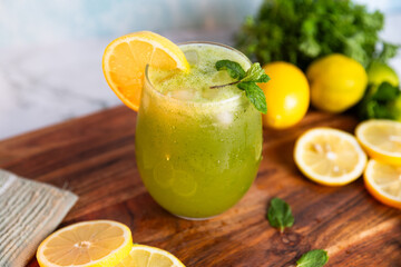 A refreshing green lemonade served in a glass with ice, garnished with mint and lemon slices, placed on a wooden board with fresh lemons and herbs in the background.
