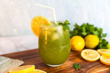 A refreshing green lemonade served in a glass with ice, garnished with mint and lemon slices, placed on a wooden board with fresh lemons and herbs in the background.