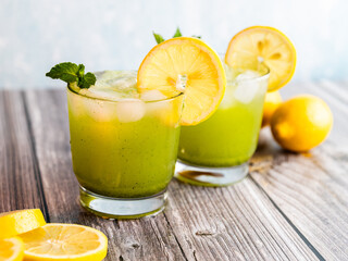 A refreshing green lemonade served in a glass with ice, garnished with mint and lemon slices, placed on a wooden board with fresh lemons and herbs in the background.