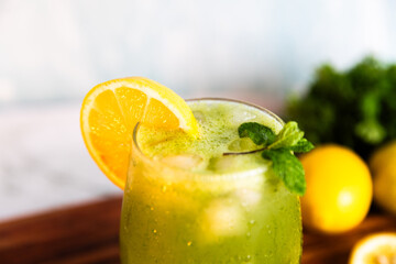 A refreshing green lemonade served in a glass with ice, garnished with mint and lemon slices, placed on a wooden board with fresh lemons and herbs in the background.