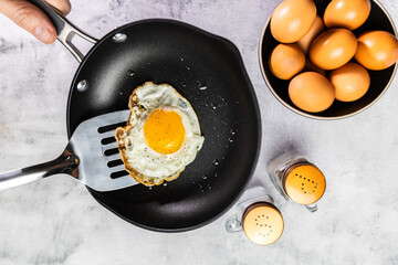 A cooked sunny-side-up egg being lifted with a spatula in a nonstick pan, with a bowl of eggs and salt and pepper shakers in the background.