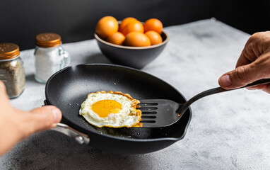 A cooked sunny-side-up egg being lifted with a spatula in a nonstick pan, with a bowl of eggs and salt and pepper shakers in the background.