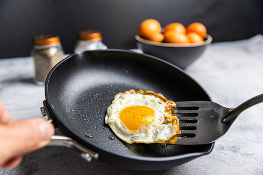 A cooked sunny-side-up egg being lifted with a spatula in a nonstick pan, with a bowl of eggs and salt and pepper shakers in the background.