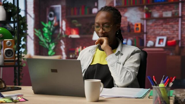 Woman in home office stretches back, feeling fatigued while working on laptop. Remote employee in apartment feeling backpain, relieving pain, solving tasks on notebook, camera B