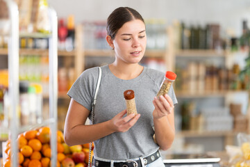 Obraz premium In healthy eating store, young woman buyer choosing spices items. Clients look package of spices bottle, inspect product and examines contents of package, compare paprika and chopped red pepper.