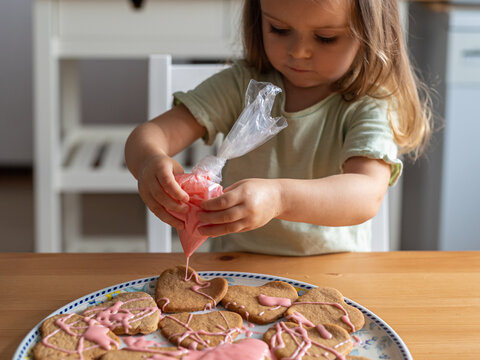 Cute little toddler girl decorating heart-shaped cookies with pink frosting