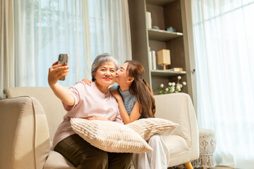 Happy Asian family elderly woman enjoy smart life technology using mobile phone at home. Healthy senior mother and adult daughter using smartphone taking selfie or vlogging together in living room. 