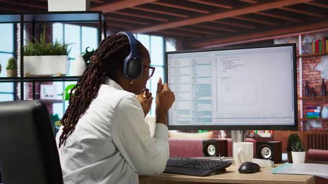 Jolly engineer in home office working on computer, dancing at desk in celebration after successfully executing code. IT worker wearing headphones happy after finishing compiling code, camera A