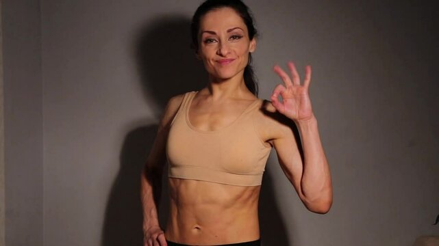 Healthy smiling woman posing and showes fingers okey sign, abdoman muscule abs, attractive body. Slim caucasian female stands in bra in studio low key light on wall home background. Sport lifestyle