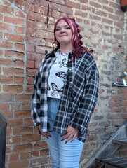 Young woman with dyed red hair wearing plaid flannel shirt posing by urban brick wall