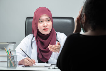 The Asian Muslim woman doctor was sitting at the patient's examination table and was examining and talking about the patient with a smiling and worried face at hospital.