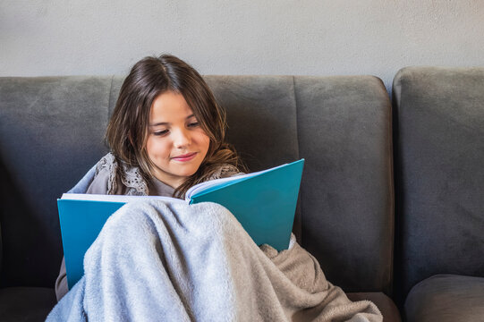 Smiling girl reading a book wrapped in a blanket on sofa