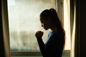 Young woman with hands folded praying to god next to window 
