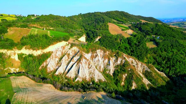 Profound aerial pullback pan left shot from Castignano (AP), the arid slopes of the badlands coming into full view, surrounded by extensive green vegetation, thriving thickets, smooth hills, blue sky