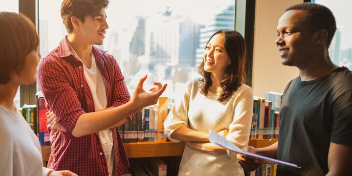 Group of diverse young adults talking, smiling, and engaging in a library setting. Diverse group enjoying conversation and discussion in a library. Diverse high school students at library.