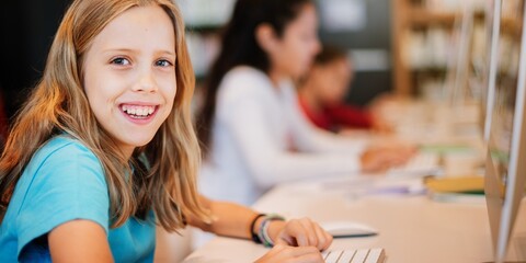 Young girl smiling at a computer in a classroom. Focused on learning, the girl is engaged in a digital activity. Classroom setting with students at computers learning with copy space.