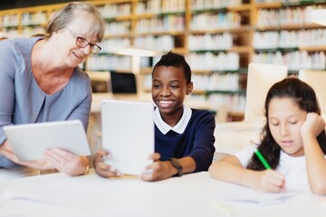 Smiling student boy uses a tablet in a library, guided by woman teacher. Diverse teacher and student hold tablets, engaging, learning activity together in the library. Teacher teaching boy in class