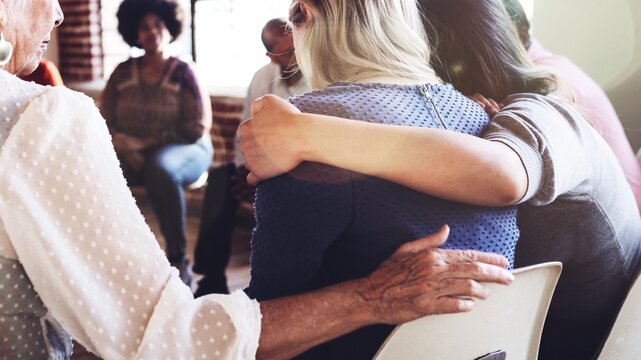 Support group meeting with diverse individuals sitting in a circle hugging and supporting one another. People offering comfort and empathy, showing support and connection in a community setting.