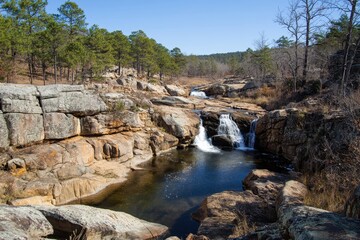 Obraz premium Scenic View of Serene Waterfalls Flowing Through Rocky Landscape Surrounded by Lush Green Trees Under Clear Blue Sky in Natural Wilderness Area