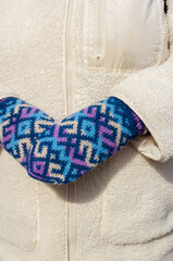 Woman dressed in beige wool coat paired with blue patterned knitted mittens. Warm and stylish outfit. Cozy, rustic charm, ideal for autumn or winter outdoor streetwear.Handmade accessories.