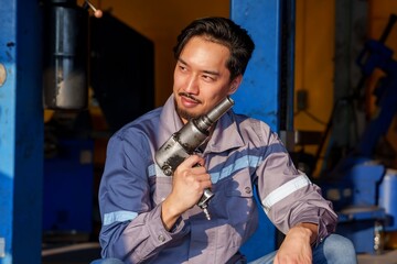 Male mechanic wearing a gray work uniform sitting while holding an air wrench tool in a garage with natural light showing confidence, expertise, and professionalism in automotive repair.