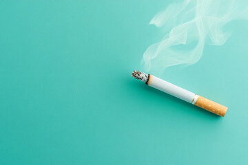 A cigarette with a brightly glowing tip and smoke curling up set against a plain blue background
