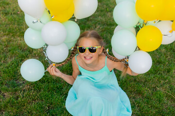 Happy little blonde girl in sunglasses and balloons on a green lawn in summer