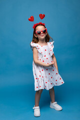 Cute little girl wearing white dress with red hearts print, red heart shaped glasses and head rim with red hearts on blue background. Happy Saint Valentine's day celebration. Birthday, Mother's day