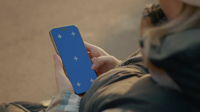 Female relaxing on outdoor bench during winter, holding smartphone with blue chroma key screen for digital communication and content mockup