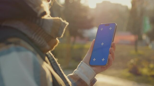 Young woman is holding a smartphone with a blue chroma key screen outdoors in a sunny day