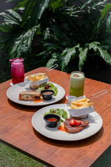 Beef Steak and Beef Ham with French Fries, Fresh Salad, and Sauces on a White Plate on a Wooden Table, Served with Dragon Fruit and Kiwi Juice.