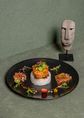 White Rice Dish Topped With Fried Egg, Accompanied By Sambal, Fresh Vegetables, And Minimalist Art Decor On A Black Plate With Decorative Statue In The Background.