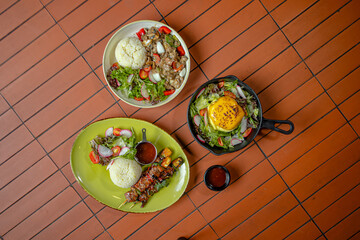 Dishes Of Steamed Rice With BBQ Beef Skewers, Creamy Mushroom Sauce, And Melted Cheese Topping, Served With Fresh Salad On A Wooden Table Viewed on Top.