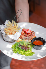 Hand Holding A Juicy Beef Steak With Fresh Salad, Savory Sauce, And French Fries, Served On A White Plate, A Rich And Flavorful Meal.