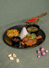 Grilled Ribs Served With Rice, Salad, A Bowl Of Soup, And Sweet Soy Sambal On A Black Plate, Garnished With Onion And Chili Around It.