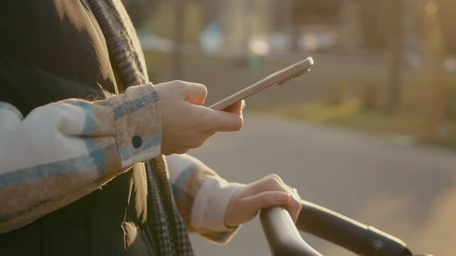 Multitasking young mother gently rocking stroller while browsing smartphone during peaceful park walk, balancing parenting and digital connection