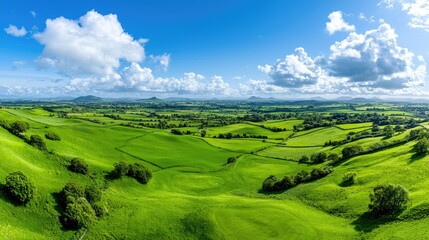 Obraz premium Lush green rolling hills stretch across a vast rural valley under a bright blue sky with fluffy white clouds during daytime.