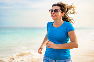 Beautiful happy middle aged woman tourist running on sandy beach on summer day. Holidays on Sal...