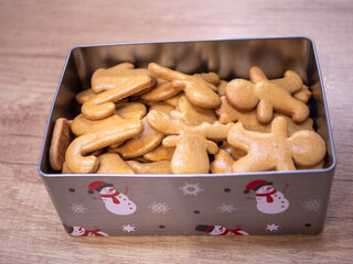 Pictured is a metal tin filled with homemade undecorated gingerbread in a variety of festive shapes such as mittens and gingerbread men. Cookies have a golden brown color and a smooth surface, placed 